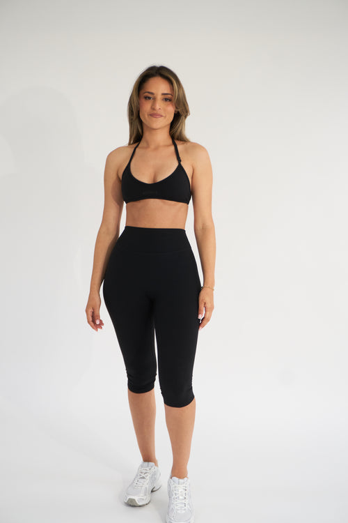 A woman modeling a black sports bra and matching black high-waisted capri leggings against a plain white studio background.