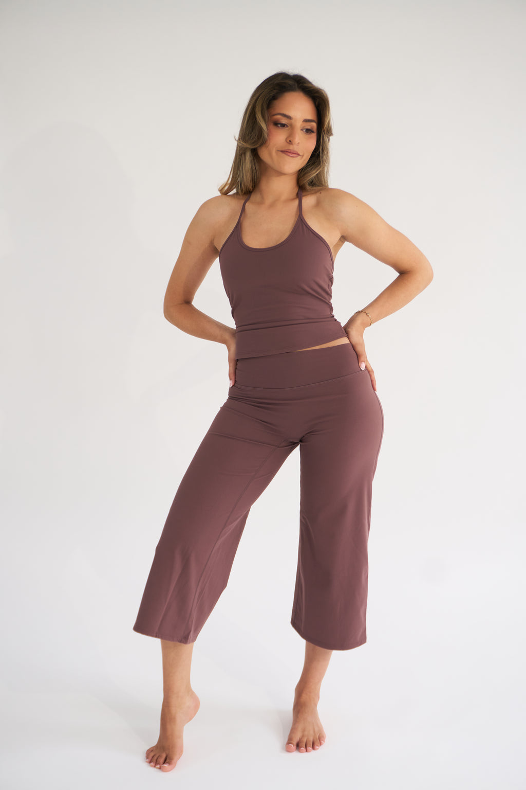 A woman modeling a matching two-piece activewear set consisting of a ribbed tank top and high-waisted cropped pants in cocoa, set against a plain white studio background.