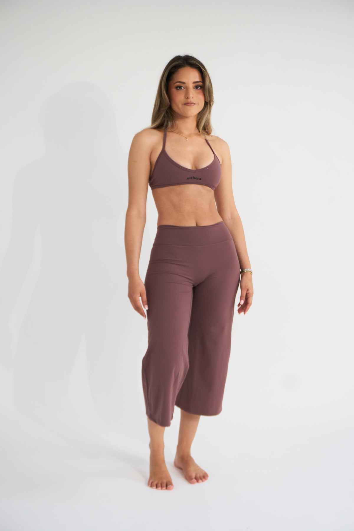 A woman modeling a matching two-piece activewear set in cocoa, consisting of a sports bra and high-waisted cropped pants, against a plain white studio background.