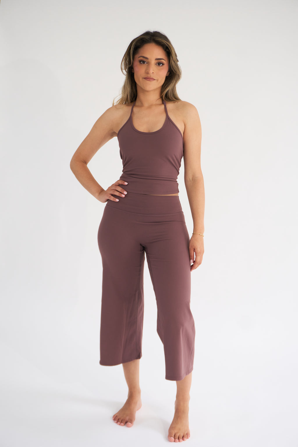 A woman modeling a matching two-piece activewear set in cocoa, consisting of a halter crop top and high-waisted cropped pants, against a plain white studio background.