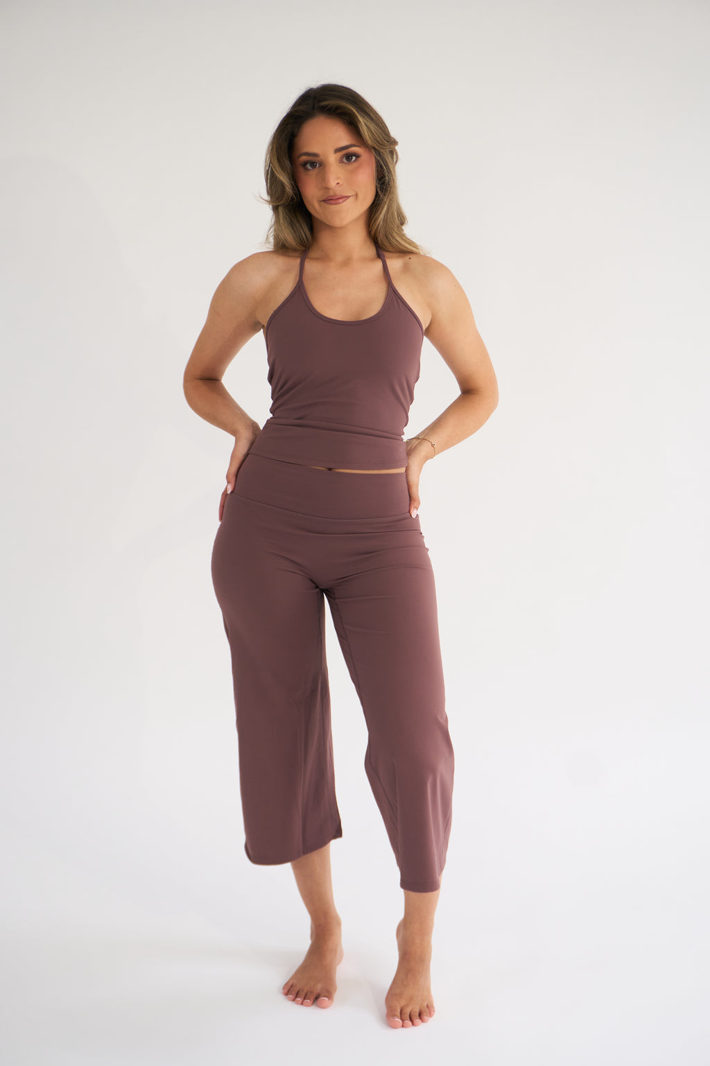 A woman modeling a matching two-piece activewear set consisting of a halter-neck tank top and high-waisted cropped pants, in cocoa, against a plain white studio background.