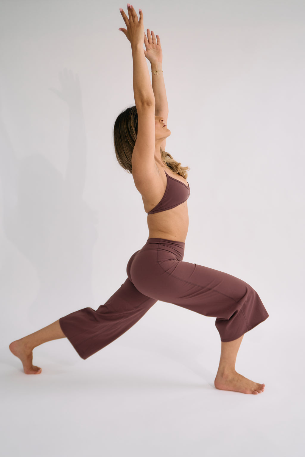 A woman in a matching cocoa activewear set poses in a yoga lunge position against a plain light gray studio background.