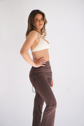 A woman modeling a cocoa two-piece activewear set consisting of a cocoa sports bra and high-waisted brown ruched flare leggings, posed against a plain light gray studio background.