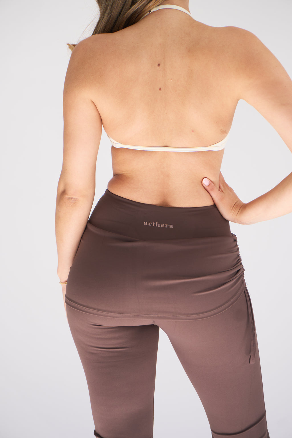 Rear view of a woman wearing high-waisted ruched cocoa leggings in a taupe brown color with 'aethera' printed on the waistband, paired with a white sports bra against a light gray background.