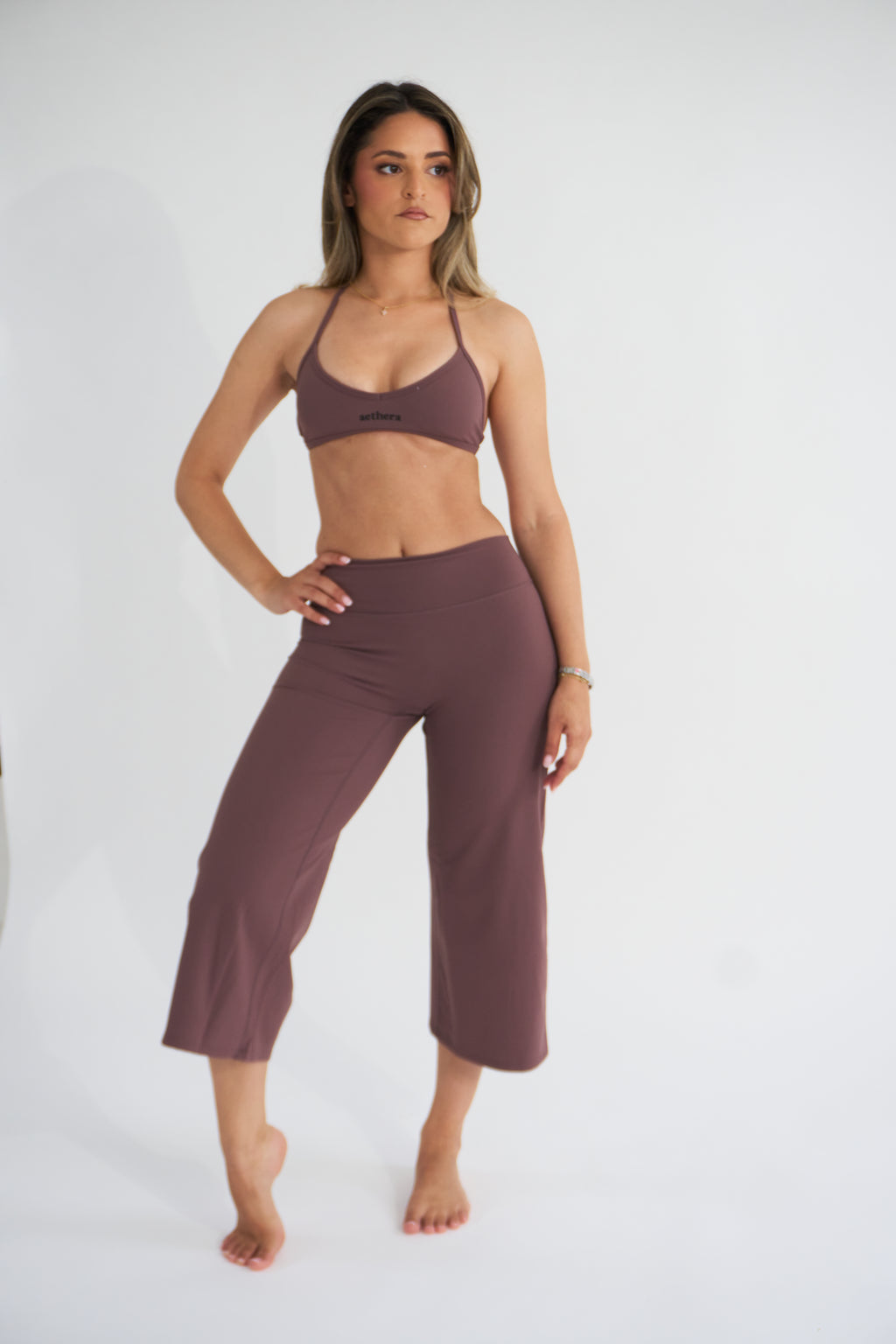 A woman modeling a matching cocoa activewear set consisting of a sports bra and cropped pants, standing against a plain white studio background.