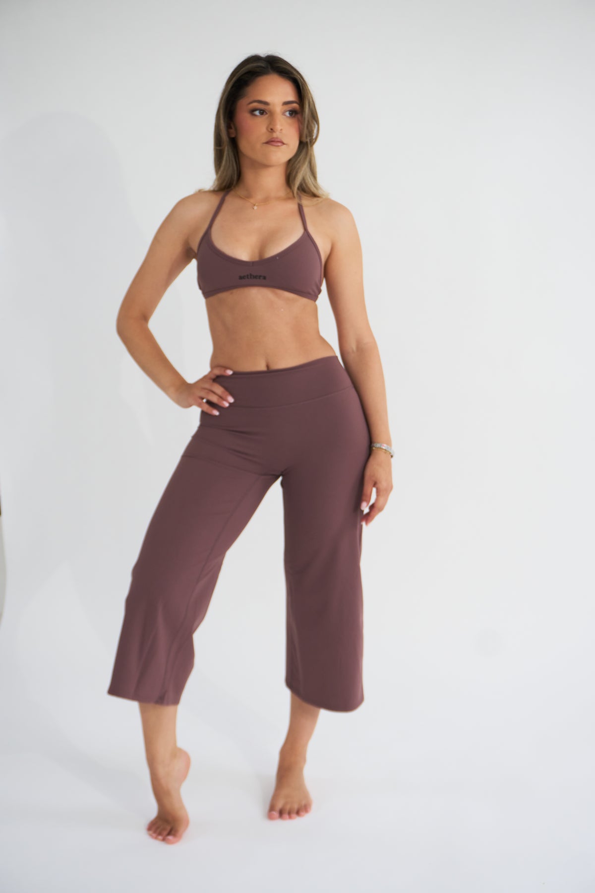 A woman modeling a matching cocoa activewear set consisting of a sports bra and cropped pants, standing against a plain white studio background.