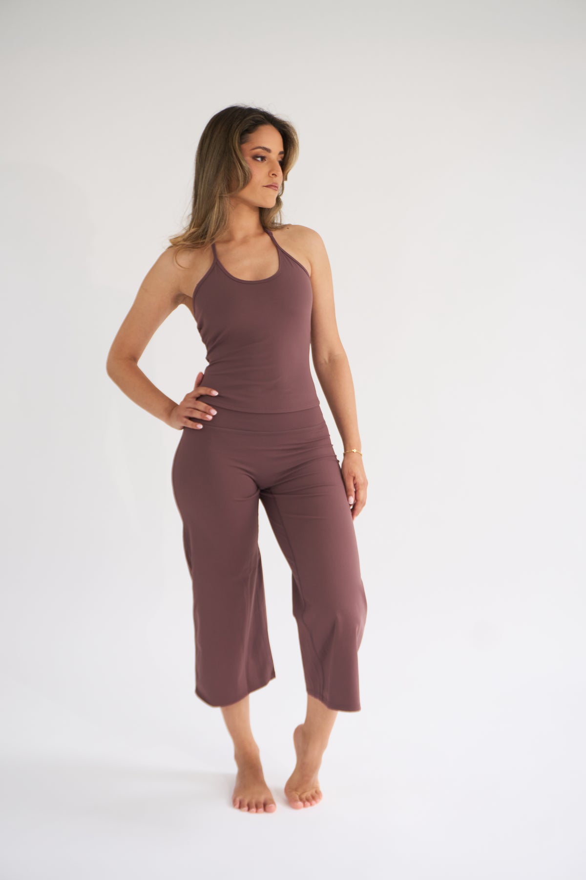 A woman modeling a matching two-piece activewear set in cocoa, consisting of a halter-neck tank top and high-waisted cropped pants, against a plain white studio background.